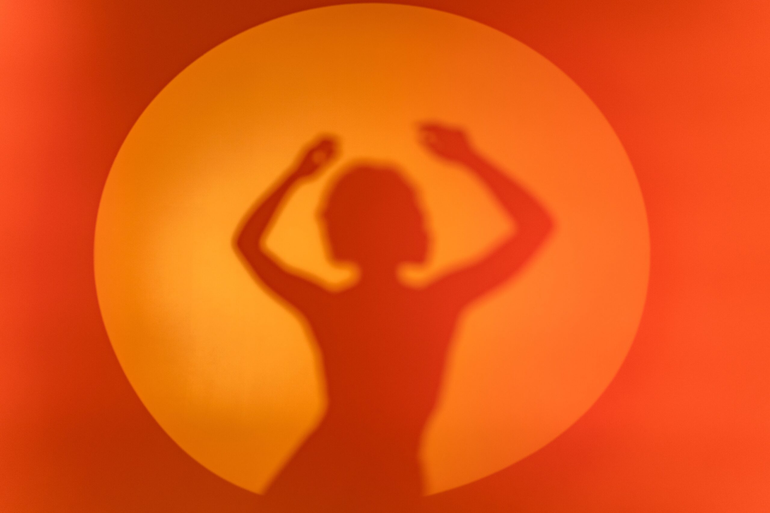 A silhouette of a woman's shadow dancing against a vibrant orange circular background.
